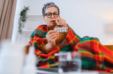 Son sınıf öğrencisi hasta bir kadın battaniyeye sarılmış, elinde ilaçlarla geziyor, evde ilaç almaya hazırlanıyor.