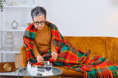 Yaşlı kadın kanepeye sarılmış, kendini iyi hissetmiyor ve evde dinlenirken grip ya da soğuk algınlığı ilacı alıyor.