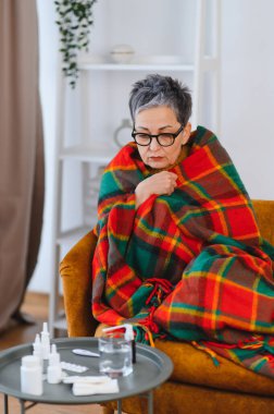 Soğuk algınlığı ya da gripten titreyen yaşlı bir kadın, sıcak bir battaniyeye sarılmış, ilaç ve suyla bir sandalyede oturuyor.