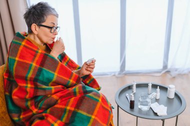 Yaşlı kadın battaniyeye sarılmış, kendini iyi hissetmiyor, ilaç alıyor ve nezle veya grip için burun spreyi kullanıyor.