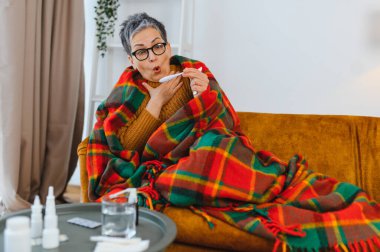 Kanepede battaniyeye sarılmış, termometreyle ateşini ölçen, ateş belirtilerine tepki veren olgun bir kadın.