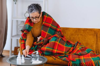 Yaşlı kadın kendini iyi hissetmiyor, battaniyeye sarılmış, ilaç ve termometreye uzanıyor.