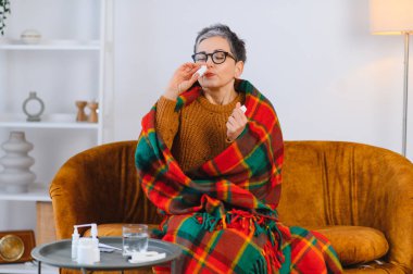 Olgun bir kadın kanepeye battaniye örterek nezle ya da grip belirtilerini azaltmak için burun damlası kullanıyor.