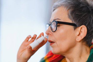 Yaşlı kadın, nezle, soğuk algınlığı ya da alerji belirtilerinden kurtulmak için burun spreyi kullanıyor.