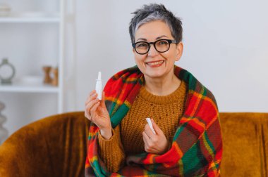 Kadın kendini daha iyi hissediyor, battaniyeye sarılmış, elinde burun spreyi ve doku tutuyor, soğuk algınlığı ve grip belirtilerinden kurtuluyor.