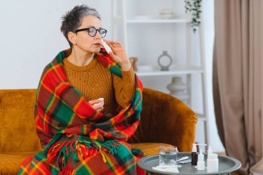 Olgun bir kadın kendini iyi hissetmiyor. Kanepede battaniyeye sarılıp grip belirtileri için burun spreyi kullanıyor. Sağlıkla ilgilenmek