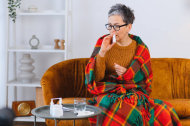 Kendini kötü hisseden bir kadın, kanepede battaniyeye sarılıp burun tıkanıklığı ve grip için dekonjestan sprey kullanıyor.
