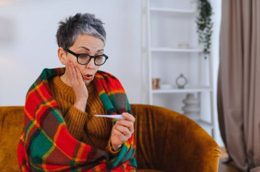 Olgun bir kadın şok olmuş bir ifadeyle termometreye bakıyor. Hastalık, ateş ya da grip belirtileri gösteriyor.