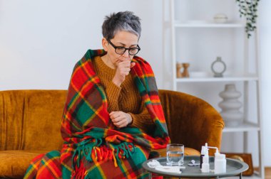 Yaşlı kadın soğuk algınlığı ya da gripten muzdarip, battaniyeye sarılı ve öksürüyor, yanında ilaçlarla.