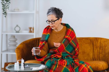 Yaşlı kadın kendini kötü hissediyor, battaniyeye sarılmış, soğuk algınlığı ve grip belirtileri için bir bardak suyla ilaç alıyor.