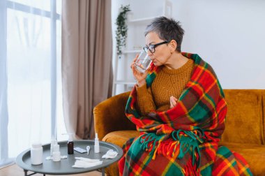 Yaşlı kadın battaniyeye sarılmış, kendini iyi hissetmiyor ve soğuk algınlığı veya grip sırasında evde dinlenirken suyla ilaç alıyor.