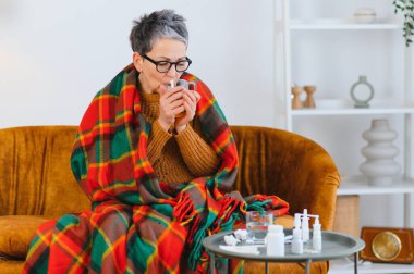 Kendini kötü hisseden bir kadın, ekose bir battaniyeye sarılmış, soğuk algınlığı veya virüsle savaşırken semptom rahatlaması için ılık bir içecek içiyor.