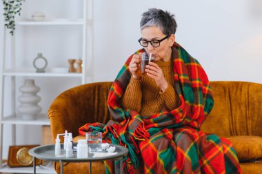 Yaşlı kadın soğuk algınlığı ya da gripten muzdarip, sıcak çay içiyor ve kanepede rahat bir battaniyenin altında ilaç içiyor.