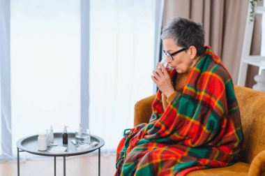 Yaşlı kadın kendini kötü hissediyor, battaniyeye sarılmış, soğuk algınlığı veya grip belirtileri için ilaç alırken su içiyor.