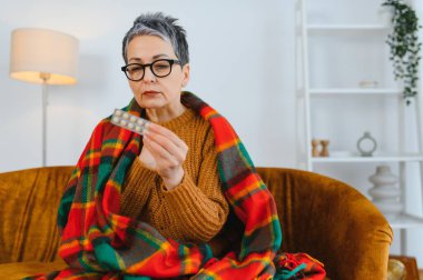 Yaşlı kadın kendini iyi hissetmiyor, battaniyeye sarılmış, soğuk algınlığı veya grip tedavisi için bir paket hap tutuyor.