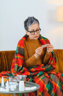 Olgun bir kadın kendini iyi hissetmiyor, battaniyeye sarılmış, elinde bir paket hapla masada ilaç tutuyor.
