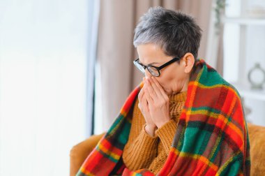 Soğuk algınlığı, grip ya da mevsimsel alerjiden muzdarip olan yaşlı bir kadın, evde hapşırırken burnunu ellerle kapatıyor.