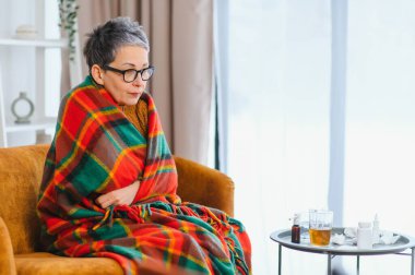 Yaşlı bir kadın, sıcak bir battaniyeye sarılmış, kanepede titriyor. Soğuk algınlığı veya grip gibi hastalık belirtileri gösteriyor.