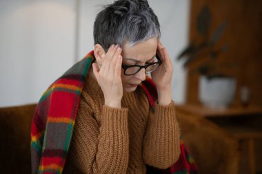 Baş ağrısı çeken, elinde şakaklar tutan ve gözlük takan bir kadın. Evde stres, hastalık ve hastalıktan muzdarip.