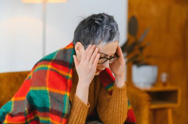 Şiddetli baş ağrısı çeken bir kadın, hastalığından ve rahatsızlığından dolayı sıcak bir battaniyeyle örtülmüş şakaklarını tutuyor.