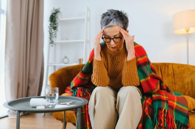 Son sınıf öğrencisi kadın yoğun baş ağrısı çekiyor. Kanepede oturuyor. Yanında battaniye ve ilaç var.