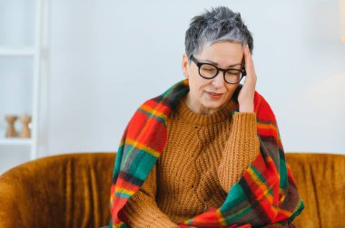Baş ağrısından muzdarip yaşlı bir kadın, sıcak bir ekose battaniyeyle örtülü, şakağını tutarken dinlenen gözleri kapalı.