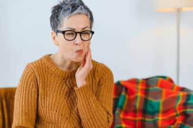 Şiddetli diş ağrısına dayanan gri saçlı olgun bir kadın. Evde yanağını rahatsızlık içinde tutuyor. Diş sağlığı ve ağrı kavramı