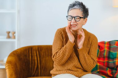 Gözlüklü olgun bir kadın diş ağrısından dolayı yoğun bir acı hisseder. Kanepede otururken yanağına dokunur.