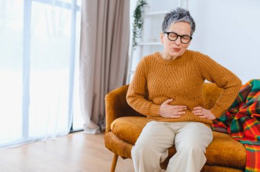 Olgun bir kadın evdeki koltukta oturuyor, şiddetli mide ağrısı ve hazımsızlıktan muzdarip, karnının üstünde el ele tutuşuyor.