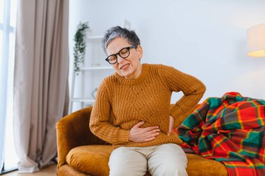 Olgun bir kadın koltukta oturup karnını kavrıyor ve sırtı ağrıyor. Sindirim güçlüğü çekiyor.
