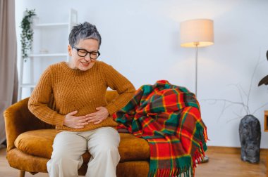 Yaşlı bir kadın ağır karın ağrısı çekiyor. Kanepede otururken karnını tutuyor. Hastalığı ve sağlık sorununu temsil eder.