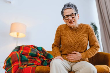 Son sınıf öğrencisi bir kadın koltukta oturuyor, karın ağrısı ve karın ağrısı çekiyor, gözleri kapalı karnını tutuyor.