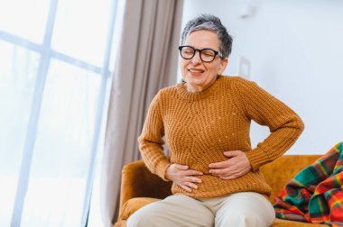 Kanepede oturan yaşlı bir kadın, karnında keskin bir ağrı ve hazımsızlık şikayetiyle karnını tutuyor.