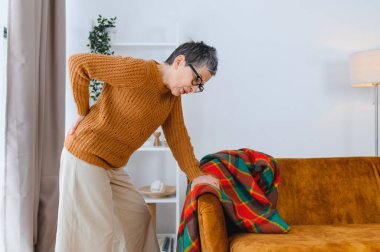 Evde acı çeken yaşlı bir kadın. Sırtını buruşturup destek için kanepeye tutunuyor.