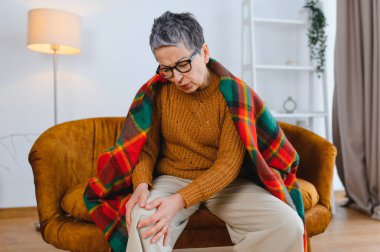 Diz eklemi ağrısından muzdarip yaşlı bir kadın, evdeki bir koltukta oturuyor, rahatsızlık yaşıyor, yaşla ilgili sağlık sorunlarını gösteriyor.