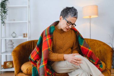 Evde diz ağrısı çeken yaşlı bir kadın, ağrıyan dizini ovuyor, rahatsızlık ve eklem iltihabı belirtileri gösteriyor.
