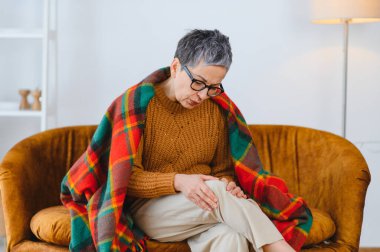 Diz eklemi ağrısından muzdarip yaşlı bir kadın, ekose bir battaniyeyle kanepede oturuyor, dizini tutuyor, rahatsızlık veriyor.