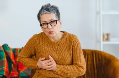 Evde keskin göğüs ağrısı çeken yaşlı bir kadın var. Elleri göğsünü sıkıyor, rahatsızlık ve sağlık sorunları gösteriyor.