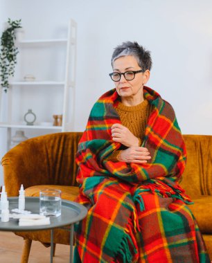 Yaşlı kadın gripten ve titremeden rahatsız oluyor. Kanepeye sarılmış, yanında ilaçlarla oturuyor.