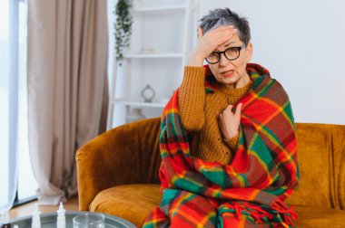 Yaşlı kadın kendini iyi hissetmiyor, sıcak bir battaniyeyle örtüyor ve baş ağrısı çekiyor, hastalık sırasında alnına dokunuyor.