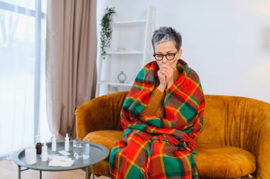 Yaşlı kadın hastalıktan muzdarip, sıcak bir battaniyeye sarılı ve kanepede otururken öksürüyor. İlaçlar masada.
