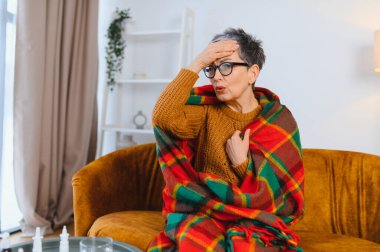 Yaşlı kadın hastalık belirtileri gösteriyor, baş ağrısı ve ateş yüzünden alnını hissediyor, kanepede battaniyeye sarılmış.