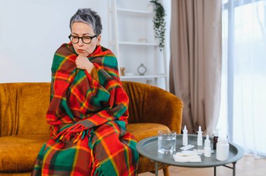 Evdeki kanepede otururken sıcak bir battaniyeye sarılmış titreyen son sınıf öğrencisi kadın, yakınlardaki bir masada birçok ilaç.