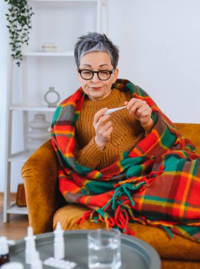 Yaşlı kadın termometreyi kontrol ediyor, evde kendini kötü hissediyor. Battaniyeye sarılmış, hastalıkla savaşıyor.
