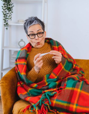 Evde battaniyeye sarılmış yaşlı bir kadın, termometreyle ateşini ölçerken yorgun ve bitkin görünüyor.