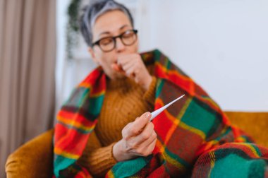 Kadın kendini iyi hissetmiyor, battaniyeye sarılmış, öksürürken termometreyle ateşini ölçüyor, hastalık ve ateşi gösteriyor.