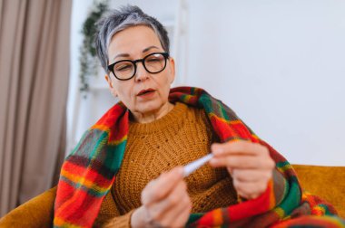 Üst düzey bir kadın battaniyeyle üstünü örtüyor termometreyi kontrol ediyor, grip belirtileri gösteriyor, ateşi var ve evde hastalık var.