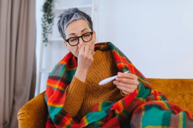 Hasta son sınıf öğrencisi bir kadın kanepede kendini kötü hissediyor. Elinde termometre var ve burnunu kapatıyor. Grip ya da soğuk algınlığı geçiriyor.