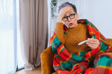 Grip, ateş, termometre tutan, battaniyeye sarılmış, baş ağrısı çeken yaşlı bir kadın.