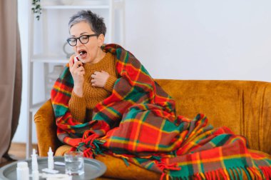 Yaşlı kadın soğuk algınlığı ya da gripten rahatsız oluyor, sıcak bir battaniyenin altında kanepede dinlenirken boğaz spreyi kullanıyor.